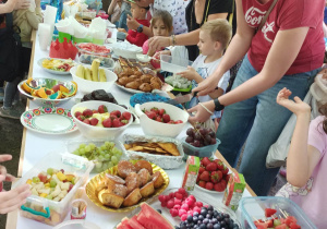 Rodzice i dzieci przy stołach, na których są kolorowe przekąski - arbuzy, borówki, winogrona, babeczki, szaszłyki owocowe