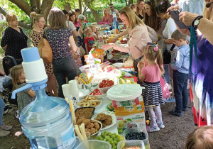 Zdjęcie prezentuje stoły z przekąskami, przy których stoją dzieci wraz z rodzinami